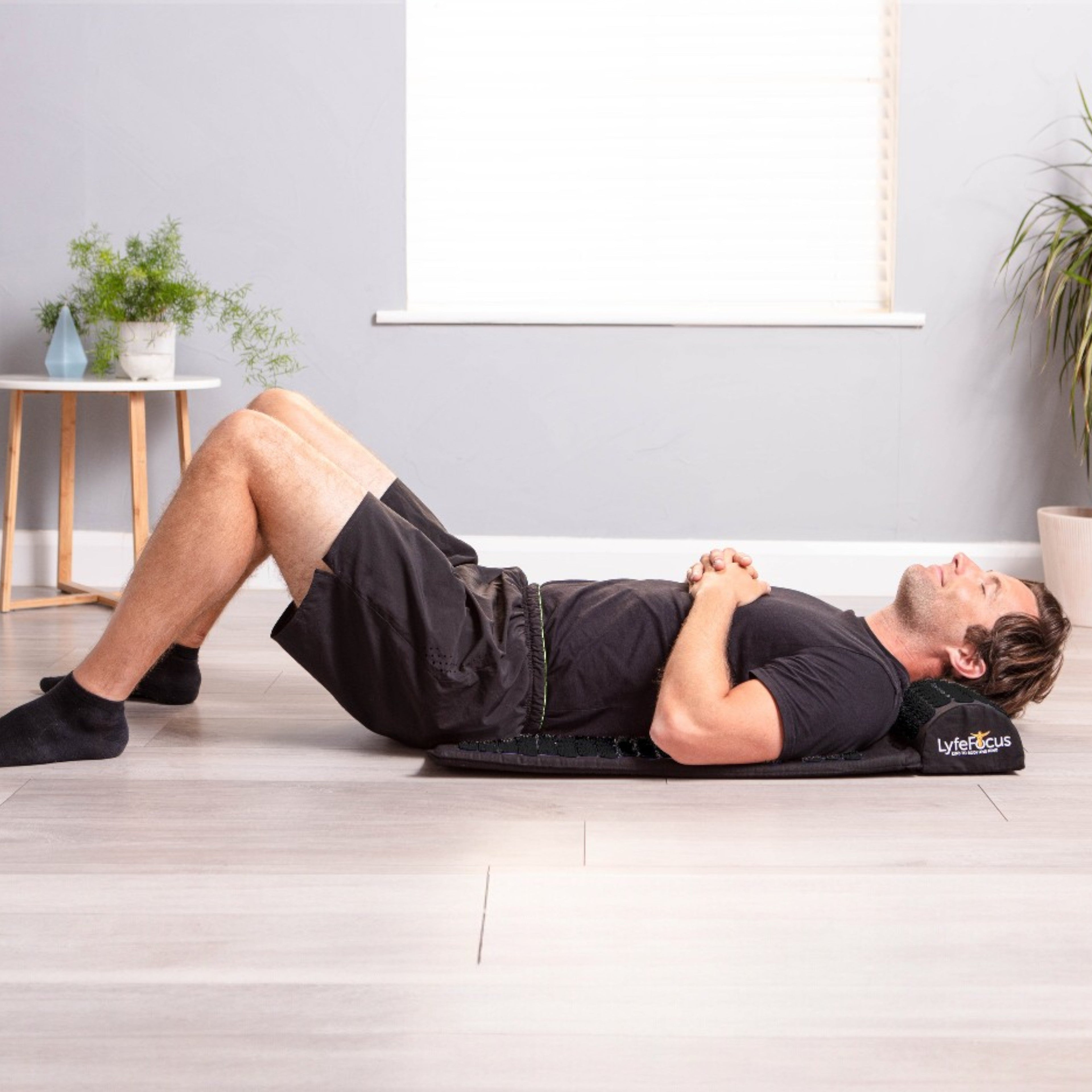 Man lying on a mat with a pillow under his head in a room with plants and a table.