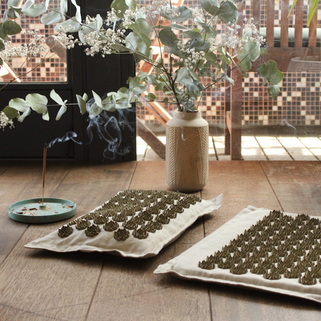 Two trays with green leafy substances on a wooden surface, with a vase of plants in the background.